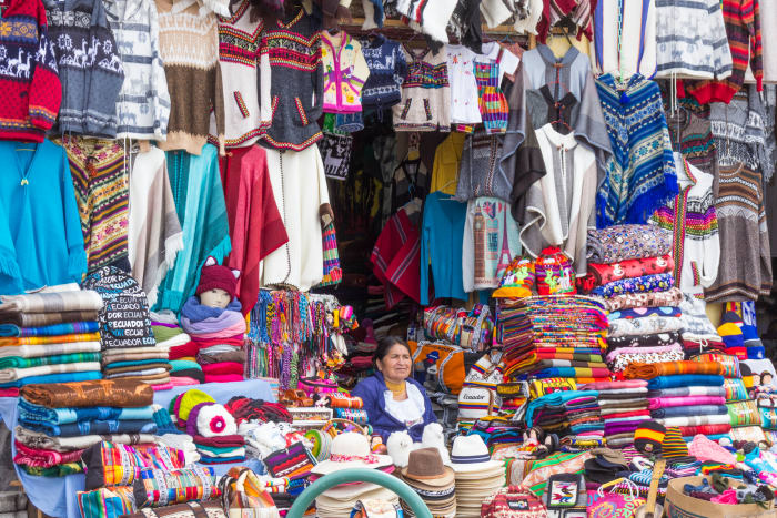 Street vendor sits at entrance to store, with wall covered in sweaters, ponchos, clothing and hats behind her