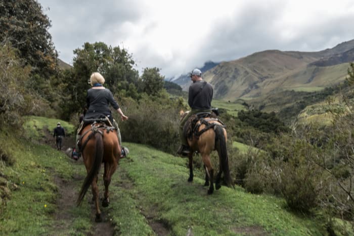 Two people on horses ride along a trail in the green mountain landscape
