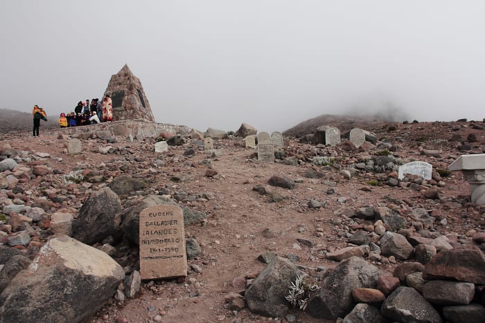 Visitors standing beside monument to climbers who died on the mountain, with gravestones on the hillside
