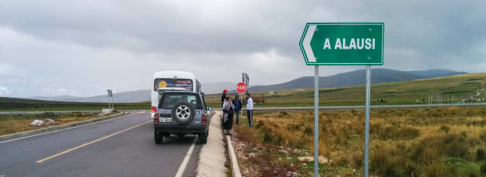 Truck and bus stopped on the side of the road, surrounded by fields, meadows and a road sign pointing to the town of Alausí