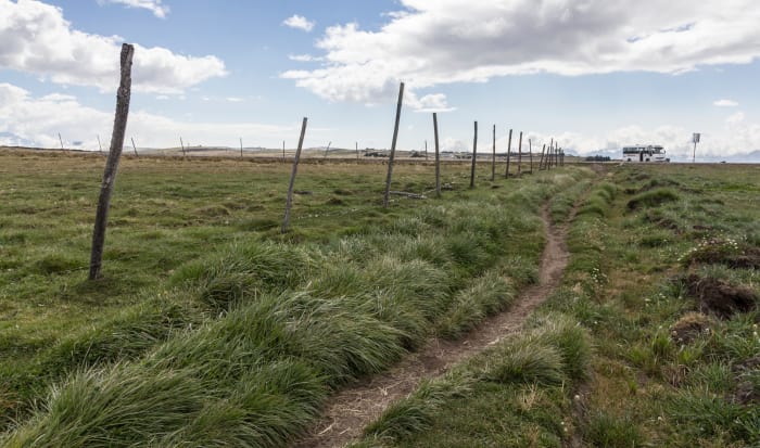 Rough dirt path in open grass field with fence posts, tour bus in distance